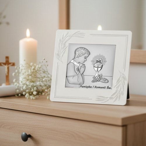 Pamiątka I Komunii Świętej – srebrny obrazek w białej ramce z liśćmi dla chłopca, aranżacja na komodzie, świece i krzyż w tle