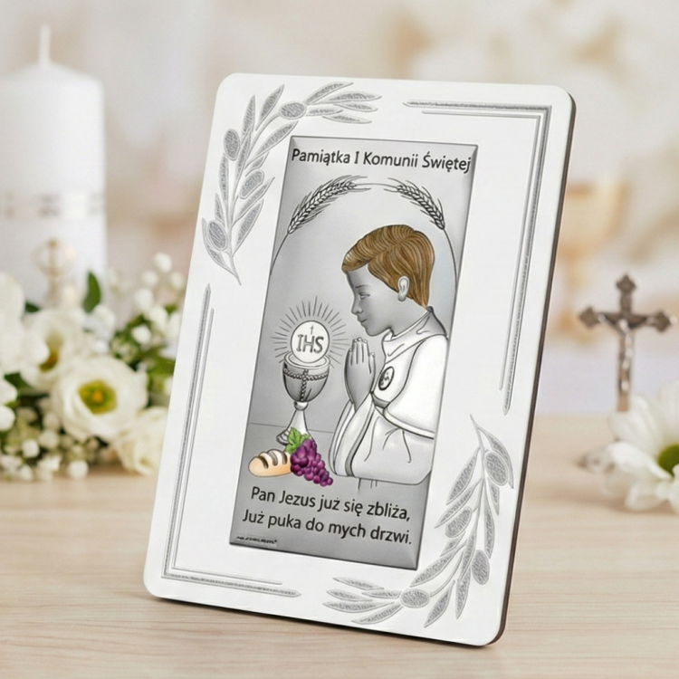 Pamiątka Pierwszej Komunii Świętej – obrazek z chłopcem w modlitwie i hostią IHS ustawiony na blacie, w tle kwiaty i krzyż