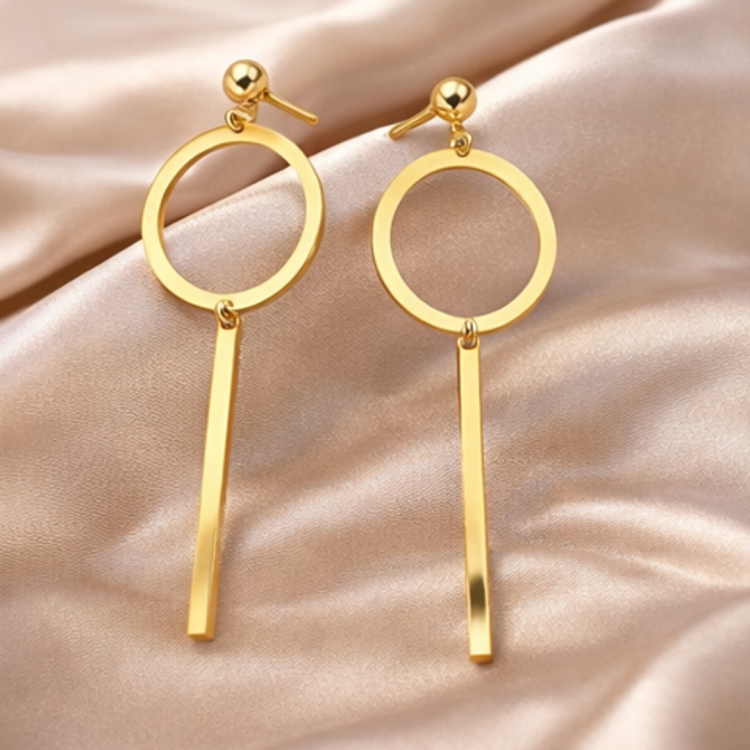 Złote kolczyki wiszące z geometrycznym okręgiem i długą pałeczką na satynowej tkaninie