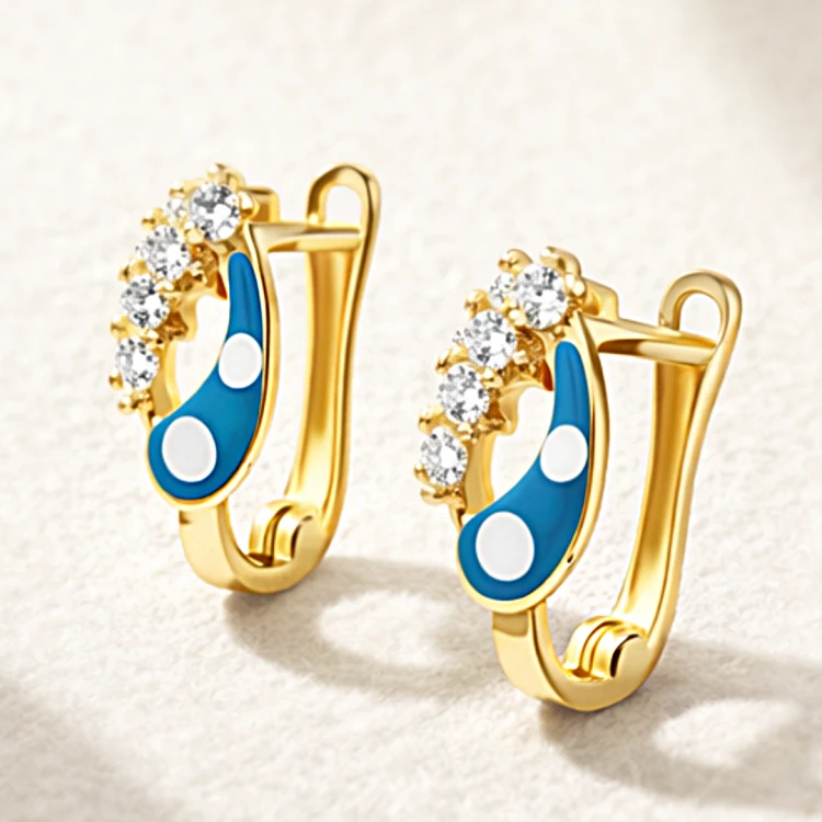 Złote kolczyki z niebieską emalią, białymi detalami i połyskującymi kamieniami na jasnym tle