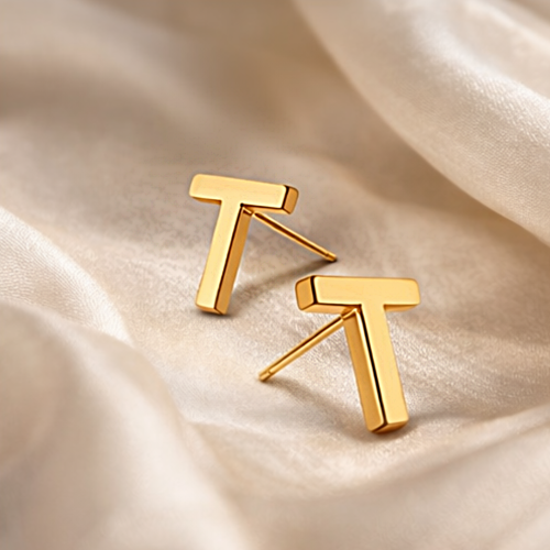 Złote kolczyki sztyfty w kształcie litery T na jasnym satynowym tle
