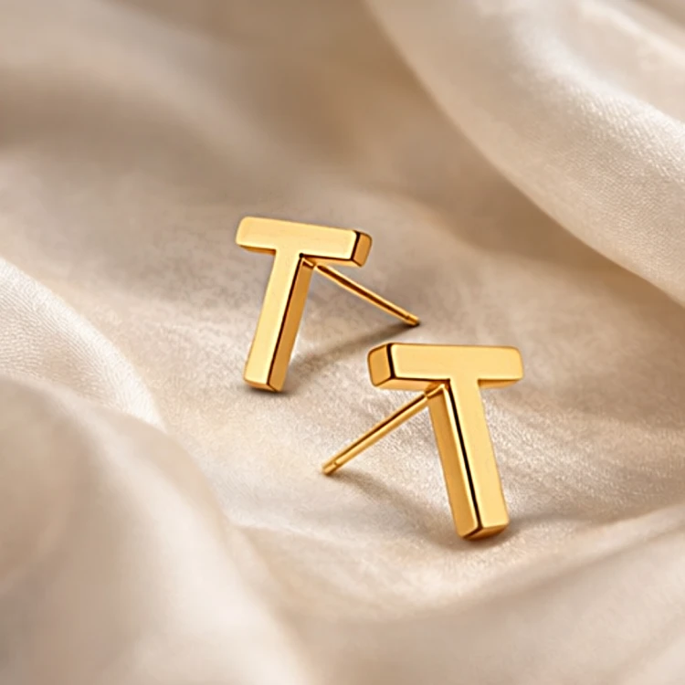 Złote kolczyki sztyfty w kształcie litery T na jasnym satynowym tle
