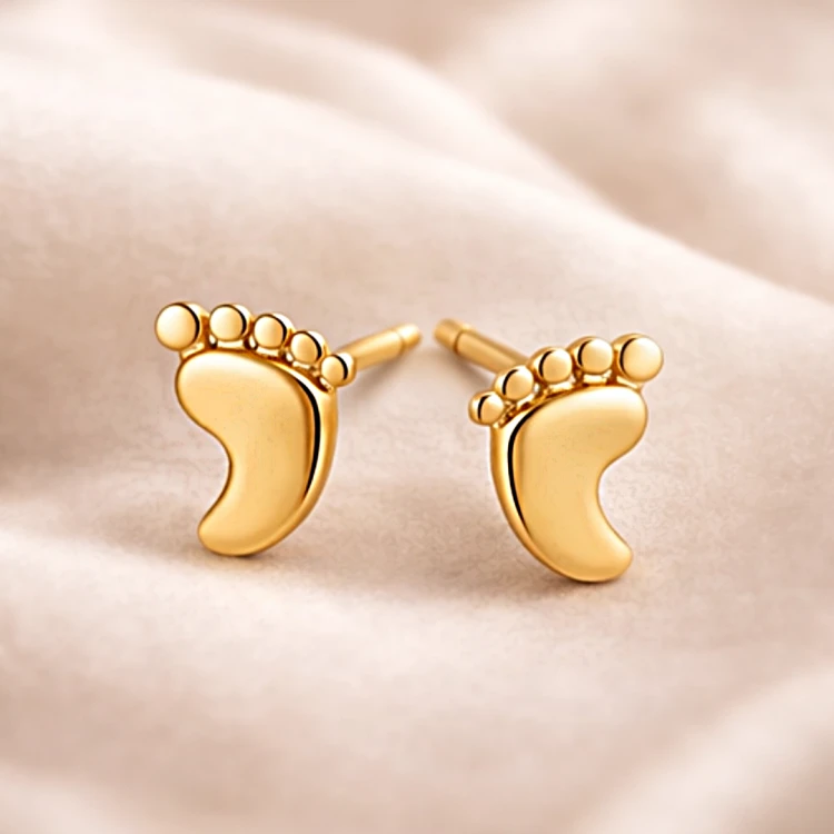 Para złotych kolczyków w kształcie stópek na jasnej tkaninie